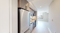 This contemporary single-floor unit showcases a clean, modern design throughout, with light hardwood-style flooring and a neutral color palette that creates a bright and cohesive feel across all spaces. The galley-style kitchen is a standout feature, equipped with stainless steel appliances including a full-size refrigerator, electric range, built-in microwave, and dishwasher. Wood-grain cabinetry with matte black hardware lines the walls from floor to ceiling, complemented by white quartz countertops and a classic white subway tile backsplash, adding texture and depth to the space. A stainless steel sink with a matte black faucet anchors the workspace. The open layout transitions seamlessly from the kitchen into the living area, which is bathed in natural light from large floor-to-ceiling windows offering expansive city views. The bedroom continues the light, airy aesthetic with a generous sliding-door closet featuring built-in shelving and hanging rods. The full bathroom is finished with large-format beige floor tiles, wood-tone vanity cabinetry with white countertops, and a tiled soaking tub and shower combination, creating a calm and functional retreat. Recessed lighting throughout adds a polished, modern touch.