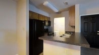 This single-floor residential space offers a straightforward and functional layout designed for comfortable everyday living. The main living area features wall-to-wall carpeting in a neutral tone, complemented by light gray and warm yellow-toned walls that create a welcoming atmosphere. A hallway extends from the living area, leading to additional rooms and providing a clear sense of the floor plan's organization. The space is illuminated by ceiling-mounted light fixtures throughout.

The kitchen is equipped with a full suite of black appliances, including a refrigerator, range, and over-the-range microwave. Warm wood-toned cabinetry lines the upper and lower walls, offering generous storage space. A peninsula with a dark laminate countertop provides additional prep and serving area, while a double-basin sink is conveniently positioned nearby. A pass-through view from the kitchen reveals a dedicated laundry area, adding to the practical functionality of the space. The textured ceilings are consistent throughout, contributing to the home's understated, utilitarian character. Overall, the space presents a clean, move-in ready environment with all essential amenities thoughtfully arranged on a single level.