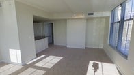 This single-floor residential unit offers a clean, open-concept layout designed for comfortable everyday living. The main living area is finished with neutral-toned wall-to-wall carpeting that adds warmth underfoot, while large windows allow natural light to pour in, casting bright patches across the floor and enhancing the airy feel of the space. Track lighting mounted on the ceiling provides adjustable illumination throughout the living area, complementing the natural light effectively. A raised architectural beam subtly defines the transition between spaces, adding a sense of structure to the open floor plan. Adjacent to the living area, the kitchen features dark granite or stone countertops set against lighter cabinetry, creating a striking contrast. The kitchen is finished with smooth tile flooring, clearly delineating it from the carpeted living space. A pass-through counter opening connects the kitchen to the main living area, promoting an open and social atmosphere. A hallway visible from the living area leads to additional rooms, suggesting a thoughtfully organized layout. The neutral color palette throughout — soft beiges and whites — creates a calm, versatile backdrop suitable for a variety of interior styles.