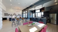 This single-floor communal lounge space offers a well-designed, open-plan layout that seamlessly integrates multiple functional zones. The interior is characterized by a two-tone color scheme, with crisp white walls contrasting against deep slate-blue accent walls, giving the space a contemporary and inviting atmosphere. Light wood-look flooring runs throughout, adding warmth to the modern aesthetic.

The space is divided into distinct yet connected areas. A dining zone features a large wooden table surrounded by bold magenta upholstered chairs, ideal for group gatherings. Adjacent to it, a wall-mounted flat-screen TV is positioned above a paneled accent wall, creating a media-focused lounge area. Further in, high-top bar tables paired with industrial-style adjustable stools provide a casual seating option.

A bar counter area lined with dark bar stools and a mounted TV overhead serves as a social hub, complete with counter space and cabinetry. Decorative wall art featuring a repeating geometric tree motif adds a distinctive visual element throughout. Recessed lighting and a chandelier fixture provide layered illumination, while large windows along the perimeter allow abundant natural light to fill the space.