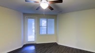 This single-floor space offers a clean, functional layout highlighted by dark hardwood-style flooring that contrasts elegantly with the light yellow-toned walls and bright white trim. The main living area is well-lit, benefiting from natural light streaming through a horizontal-blind-covered window and a glass-panel exterior door, which also provides direct outdoor access. A ceiling fan with an integrated light fixture is centrally positioned, ensuring year-round comfort and overhead illumination. The walls are smooth and neutral, offering a versatile backdrop suitable for a variety of interior styles. Recessed HVAC vents are integrated into the ceiling, contributing to a tidy, uncluttered aesthetic. The space feels open and airy, with a straightforward design that emphasizes practicality. The combination of durable flooring, ample natural light, and climate control features makes this a comfortable and adaptable living environment.