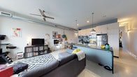This single-floor space showcases an open-concept layout that seamlessly connects the living, dining, and kitchen areas into one cohesive environment. The living area is anchored by a large sectional sofa paired with a bold geometric area rug, a media console with shelving, and a ceiling fan with exposed ductwork overhead—lending an industrial character to the space. Recessed and pendant lighting throughout provides both functional illumination and visual interest.

The kitchen is positioned along one side of the open floor plan, separated from the living area by a dark-countertop breakfast bar that doubles as a casual dining surface. Light wood cabinetry lines the upper walls, complemented by stainless steel appliances including a refrigerator, microwave, and range. Industrial-style pendant lights hang above the counter, reinforcing the modern aesthetic.

A compact dining area with a small table and stools sits between the living and kitchen zones, making efficient use of the available space. The light-toned flooring and neutral wall colors contribute to a bright, airy atmosphere, while pops of yellow and greenery add warmth and personality throughout.