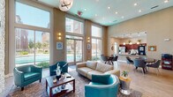 This single-story community center in Baton Rouge, Louisiana, is designed with an open, airy layout highlighted by soaring ceilings that lend a grand sense of scale throughout the space. The main lounge area serves as the centerpiece, featuring a striking floor-to-ceiling stacked stone accent wall with a built-in fireplace and a mounted flat-screen television. Teal leather club chairs and a cream leather sofa are arranged around a wood coffee table atop a decorative area rug, creating an inviting and cohesive seating arrangement.

Large floor-to-ceiling windows and glass doors line one wall of the lounge, flooding the interior with abundant natural light and offering direct views of the outdoor pool area. Recessed lighting and a modern statement chandelier complement the natural light throughout.

Adjacent to the lounge, a dedicated café-style area houses a Starbucks coffee station and features light wood-look flooring, a round dining table with modern chairs, and a continuous bar-height counter along the wall fitted with stylish wood-and-chrome barstools. Pass-through windows connect this area to neighboring spaces, enhancing the open flow.

A kitchenette with pendant lighting, dark cabinetry, and stainless steel appliances is also visible, providing additional functionality. The overall ambiance is modern and welcoming, combining warm neutral tones with bold teal accents.