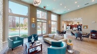 This single-story community center in Baton Rouge, Louisiana, is designed with an open, airy layout highlighted by soaring ceilings that lend a grand sense of scale throughout the space. The main lounge area serves as the centerpiece, featuring a striking floor-to-ceiling stacked stone accent wall with a built-in fireplace and a mounted flat-screen television. Teal leather club chairs and a cream leather sofa are arranged around a wood coffee table atop a decorative area rug, creating an inviting and cohesive seating arrangement.

Large floor-to-ceiling windows and glass doors line one wall of the lounge, flooding the interior with abundant natural light and offering direct views of the outdoor pool area. Recessed lighting and a modern statement chandelier complement the natural light throughout.

Adjacent to the lounge, a dedicated café-style area houses a Starbucks coffee station and features light wood-look flooring, a round dining table with modern chairs, and a continuous bar-height counter along the wall fitted with stylish wood-and-chrome barstools. Pass-through windows connect this area to neighboring spaces, enhancing the open flow.

A kitchenette with pendant lighting, dark cabinetry, and stainless steel appliances is also visible, providing additional functionality. The overall ambiance is modern and welcoming, combining warm neutral tones with bold teal accents.