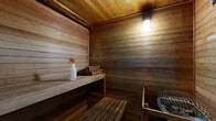 This traditional sauna is a compact, self-contained space constructed entirely with horizontal wood paneling across the walls, ceiling, and floor, creating a warm, enveloping aesthetic characteristic of classic sauna design. The interior features two tiers of wooden benches — a lower bench for seating and a raised upper bench for those seeking more intense heat — both crafted from smooth, natural wood planking. A sauna heater with a rounded metal grate sits in the corner, serving as the functional centerpiece of the space and the source of heat for the room. Soft, warm artificial lighting mounted on the upper wall casts a gentle glow throughout the interior, enhancing the intimate and calming atmosphere. A framed window on one wall looks out into an adjacent brighter room, providing a visual connection to the exterior area while maintaining the insulated environment within. A safety notice is posted near the window, indicating guidelines for safe use of the sauna. The door opens directly to what appears to be a common area with carpeted flooring. The overall ambiance is rustic and functional, offering a quiet, enclosed retreat purpose-built for heat therapy and relaxation.