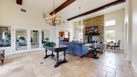 Located at 606 S Riverside Ave in Rialto, California, this single-floor community facility offers a thoughtfully designed layout encompassing a living room, exercise room, kitchen, and office. The centerpiece of the space is the expansive living room, distinguished by soaring ceilings adorned with rich dark wood beam accents and two ornate chandelier fixtures that cast a warm, welcoming glow. A dramatic floor-to-ceiling stacked stone fireplace serves as a striking focal point, complemented by plush seating arrangements and travertine tile flooring that extends throughout the space. Natural light filters in through multiple windows, enhancing the open and airy atmosphere. The kitchen features warm-toned cabinetry, granite countertops, a granite tile backsplash, and travertine flooring, providing a fully functional and attractive space. The office is outfitted with built-in cabinetry and granite-topped workstations, offering a practical and organized workspace. The exercise room is well-equipped with Matrix cardio machines, a cable weight system, a dumbbell rack, and floor-to-ceiling mirrors that amplify the sense of space, all set on durable carpeted flooring with recessed lighting overhead.