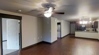 This single-floor space offers a straightforward, functional layout designed for comfortable everyday living. The main living area is open and airy, featuring rich dark hardwood-style flooring that runs throughout, providing a warm contrast against the light neutral-toned walls. A five-blade ceiling fan with an integrated light fixture serves as a central design element, offering both comfort and ambient lighting to the space. Dark baseboards provide a clean, defined border between the walls and floors, adding a subtle decorative touch.

Toward the rear of the main living area, a dedicated laundry alcove is visible, housing a stacked washer and dryer unit — a convenient in-unit feature. Beyond the living space, a hallway leads to additional rooms, with what appears to be a bedroom or closet area featuring louvered doors offering privacy and ventilation. A partial view of a kitchen counter is visible to one side, suggesting a practical kitchen space adjacent to the main living area.

The overall ambiance of the space is clean, minimal, and move-in ready, with a neutral color palette that provides a versatile backdrop for a variety of furnishing styles. The open flow between areas makes the space feel connected and easy to navigate.
