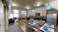 This two-floor space offers a thoughtfully designed layout that blends functionality with modern style. The heart of the home is an expansive open-concept kitchen and living area, seamlessly connected to encourage comfortable everyday living and entertaining. The kitchen is outfitted with light maple cabinetry, dark granite-style countertops, and stainless steel appliances including a range and microwave. A large central island with seating provides additional workspace and a casual dining option, while striking teal pendant lights add a vibrant pop of color against the neutral palette. Tile flooring in the kitchen transitions into carpeted living areas, creating distinct zones within the open floor plan. Tall windows along the exterior walls flood the space with abundant natural light, enhancing the airy, open atmosphere. A dedicated dining area adjacent to the kitchen features a full-sized table with seating for multiple guests. The living room area is visible from the kitchen, reinforcing the cohesive, connected feel of the main level. With two floors, the space provides ample room for both shared living and private retreats, making it a well-rounded and versatile environment suited for a variety of lifestyles.