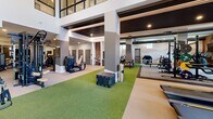 Located at 4900 East Deer Valley Drive in Phoenix, AZ, this expansive fitness center offers a generously sized, single-floor exercise facility designed to support a full spectrum of workout needs. The space features soaring ceilings in key zones, creating an open and energizing atmosphere throughout.

The cardio area is equipped with a long row of treadmills and additional cardio machines positioned alongside floor-to-ceiling windows that frame views of the outdoor palm-lined surroundings, allowing natural light to flood the space. Multiple flat-screen TVs and digital display boards are mounted throughout for entertainment and performance tracking.

The strength training zones are thoughtfully segmented, with dedicated areas for selectorized machines, free weights, and multi-functional cable systems. A comprehensive dumbbell rack and wall-length mirrors — including a motivational "No Pain No Gain" neon sign — enhance the weight room section. Rubber flooring tiles provide a durable, safe surface in the heavy lifting area.

Artificial turf runs through a central functional training corridor, accommodating activities such as sled pushes, agility drills, and bodyweight exercises. Plyo boxes and TRX suspension systems add to the functional fitness offerings. A distinct section with a decorative slatted ceiling, warm wood tones, and a scenic mural accent wall adds architectural character. The facility also includes a dedicated area with leg press and plate-loaded equipment, offering a complete, well-appointed training environment.