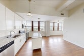 Located at 20 Beacon Way in Jersey City, NJ, this one-bedroom, one-bathroom residence offers a thoughtfully designed layout across a single floor. The open-concept kitchen and living area serves as the heart of the home, showcasing soaring ceilings with exposed wooden beams and industrial-style ductwork that lend a loft-inspired character. Rich, dark hardwood flooring runs throughout, complementing the kitchen's deep mahogany cabinetry, granite countertops, and a matching center island with pendant lighting above. Stainless steel appliances complete the kitchen's modern, functional aesthetic. A hallway connects the main living area to the private spaces, featuring the same warm hardwood flooring and whitewashed beam ceiling details. The bedroom is carpeted for comfort and benefits from generous natural light through large windows fitted with blinds, offering impressive elevated views of the Jersey City skyline and waterfront. The bathroom features neutral travertine-style tile flooring and a window that frames urban cityscape views, adding brightness to the space. Clean white walls throughout the residence create a bright, neutral canvas, while the overall design balances industrial character with modern residential comfort.