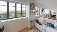 This well-appointed single-floor residence showcases a clean, contemporary design throughout. The kitchen is outfitted with high-gloss white cabinetry extending to the ceiling, stainless steel appliances including a refrigerator, range, and dishwasher, a subway tile backsplash, and light wood flooring that continues through much of the space.

The living area is bathed in natural light courtesy of large, dark-framed windows that frame sweeping views of surrounding greenery. The open layout provides a comfortable and airy atmosphere, with warm wood flooring adding to the inviting ambiance.

The residence features multiple bedrooms, each offering natural light through well-placed windows with outdoor views. The primary bedroom includes a walk-in closet with built-in shelving and hanging storage, while additional bedrooms provide generous space and a connection to the living area.

Two full bathrooms serve the residence, both finished consistently with white vanity cabinetry, undermount sinks, backlit mirrors, and distinctive hexagonal tile flooring. One bathroom includes a full tub-shower combination, while the other is configured as a half bath. Throughout, the design emphasizes a cohesive, modern aesthetic with neutral tones, clean lines, and quality finishes.