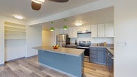 This single-floor residence offers a well-designed layout with distinct living, sleeping, and kitchen zones connected by light wood-style flooring throughout the main areas. The kitchen is a standout feature, showcasing a two-tone cabinetry design with white upper cabinets and slate-gray lower cabinets topped with neutral countertops. Stainless steel appliances — including a refrigerator, electric range, and dishwasher — complement the modern aesthetic. A central island provides additional prep and seating space, while vibrant green pendant lights add a bold, contemporary accent. Natural light enters through a window above the sink, offering a pleasant view of surrounding greenery.

The spacious living area is finished with light wood-style flooring and features multiple large windows that frame lush tree views, creating a bright and open atmosphere. Baseboard heating runs along the walls throughout. The bedrooms are carpeted for comfort, with one featuring a closet and direct access to a bathroom. The bathroom includes a soaking tub with a patterned shower curtain, a vanity with an integrated sink, a mirrored medicine cabinet, and clean, neutral finishes. The overall ambiance is clean, functional, and modern.
