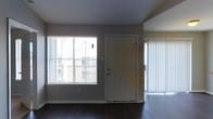 This single-floor space offers a clean, functional layout with a welcoming open-plan living area at its core. Dark hardwood floors run throughout, providing a rich contrast against the neutral gray walls and white trim, lending the space a modern yet understated aesthetic.

The main living area is brightened by a large window fitted with horizontal blinds, allowing natural light to filter in and illuminate the interior. A classic six-panel white front entry door serves as the primary access point and is flanked by the window, creating a well-composed entryway wall. On the opposite side of the space, a floor-to-ceiling vertical blind treatment covers what appears to be a sliding glass door, offering both privacy and access to an outdoor area beyond.

The ceilings are smooth and painted a cool gray tone, contributing to the calm, neutral palette maintained throughout. The overall ambiance is clean and move-in ready, with a minimalist character that allows for versatile furnishing and personalization. The abundance of natural light from multiple sources keeps the space feeling open and airy, making it both practical and comfortable as a living environment.