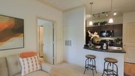 This single-story space showcases a thoughtfully designed open-concept layout that integrates the living and kitchen areas into one cohesive environment. The living area features plush carpeting and is furnished with a comfortable sofa accented by decorative pillows, creating a relaxed and welcoming atmosphere. Crown molding along the upper walls adds a refined architectural touch throughout.

The kitchen is positioned adjacent to the living space and is defined by a raised dark-toned breakfast bar, ideal for casual dining or entertaining. Dark cabinetry reaches toward the ceiling, providing generous storage, while black appliances contribute a sleek, modern contrast to the light-colored walls. A stylish pendant light and a track lighting fixture illuminate the kitchen area effectively, enhancing both functionality and ambiance.

A wall-mounted ventilation grille is neatly integrated into the shared wall between the two spaces. A white interior door with clean panel detailing leads to an additional room, visible in the background as a hallway or bathroom area. The overall palette of warm whites and neutral tones, combined with the dark kitchen accents, gives the space a balanced and modern aesthetic.