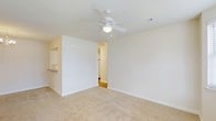 Located at 100 Millspring Dr in Durham, North Carolina, this single-floor residence offers a clean, functional layout designed for comfortable everyday living. The main living area is bright and open, featuring neutral-toned carpet flooring and white walls that create a light, airy atmosphere. A ceiling fan with an integrated light fixture is centrally positioned overhead, providing both air circulation and ambient lighting. Smooth white ceilings and crisp white baseboards give the space a tidy, well-maintained appearance throughout. An open passageway connects the living area to additional rooms, allowing for a natural flow between spaces. Adjacent to the main living area, a partially visible kitchen area features cabinetry and a countertop, suggesting a practical and accessible kitchen layout. The overall design of the residence is straightforward and clean, emphasizing usability and ease of movement across the single floor. The neutral color palette and consistent flooring choices contribute to a cohesive and welcoming interior environment, making this a well-suited space for a range of living arrangements.