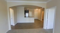 This single-story space showcases a well-designed open-concept floor plan that seamlessly connects the main living areas. A defining architectural feature is the elegant arched passageway, framed with crown molding, that links the carpeted living area to the kitchen and beyond, adding a refined visual element to the layout.

The kitchen is positioned centrally and features dark cabinetry, stainless steel appliances including a full-size refrigerator, and a peninsula with a light-toned countertop and an integrated sink. Track lighting overhead provides functional illumination, while the peninsula creates a natural division between the kitchen and the surrounding living spaces.

The flooring transitions from plush carpet in the living area to dark hardwood-style flooring in the kitchen and connecting hallways, adding both visual contrast and practicality. Crown molding runs throughout, lending a cohesive and polished finish to the neutral-toned walls.

Beyond the kitchen, the layout continues with additional doorways suggesting further rooms, offering a functional flow throughout the space. The overall ambiance is clean, open, and move-in ready.