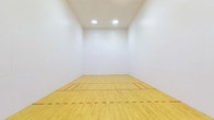 This single-level athletic facility centers around a full racquetball court designed to meet regulation standards. The court features a smooth, light-toned hardwood floor finished to a high sheen, with clearly marked red boundary and service lines that delineate the playing zones precisely. The surrounding walls are finished in bright white, providing a clean, unobstructed surface essential for gameplay, while the seamless corners and flush wall construction ensure consistent ball rebound throughout.

Overhead, recessed flush-panel lighting fixtures are evenly distributed across the ceiling, delivering uniform, shadow-free illumination across the entire court surface. The ceiling height is generously tall, accommodating the full vertical range of play expected in racquetball.

At the rear of the court, a full-width glass wall with a glass entry door separates the court from an adjoining fitness center. This transparent partition allows natural visibility between the two spaces while maintaining acoustic and spatial separation. The fitness center beyond is equipped with a range of strength and cardio machines and features vibrant yellow accent walls, additional natural light from exterior windows, and its own ventilation system.

The overall ambiance is clean, functional, and purpose-built, making this facility well-suited for both competitive play and recreational athletic use.