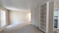 This single-floor residence offers a functional and well-organized layout across several distinct living areas. The spacious living room is finished with plush carpeting and neutral beige walls, creating a warm and inviting atmosphere. A standout feature of the space is the built-in white shelving unit that divides the room, providing generous storage and display capacity while adding architectural interest. A ceiling fan overhead and a window with vertical blinds allow for natural light and air circulation.

The galley-style kitchen is efficiently designed with gray cabinetry, dark countertops, and a stone mosaic tile backsplash. Stainless steel appliances, including a refrigerator, electric range, and dishwasher, are integrated throughout. Wood-look flooring runs the length of the kitchen, complementing the contemporary finish selections.

The bedrooms are carpeted and bathed in natural light through windows fitted with horizontal blinds. One bedroom includes a ceiling fan, while another features a pair of symmetrically placed windows. The bathroom offers wood-look flooring, a combined tub and shower, a white vanity with dark countertop, and a window providing natural ventilation. Textured ceilings are consistent throughout the unit.