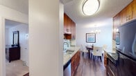 This single-floor residence offers a thoughtfully designed layout that connects living, dining, and kitchen spaces in a cohesive, functional flow. The kitchen is outfitted with rich warm-toned cabinetry, granite-style countertops, a mosaic tile backsplash, stainless steel sink, and black appliances including a range, microwave, and refrigerator. It opens directly to a dedicated dining area, which features wood-look flooring, a round dining table with upholstered chairs, a stylish chandelier, and a window that brings in natural light. The living room is carpeted and generously sized, with large sliding glass doors that open to a private balcony overlooking mature trees, providing a serene outdoor connection. The bedroom is a quiet retreat with carpet flooring, natural light through a window, a built-in closet with shelving, and ample space for a dresser and nightstands. The bathroom is clean and modern, featuring a tub-shower combo with sliding glass doors, a granite-topped vanity with warm wood cabinetry, and wood-look flooring. Throughout the residence, textured ceilings and neutral wall tones create a consistent and comfortable atmosphere well-suited for everyday living.