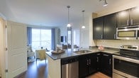 This single-floor residence offers a thoughtfully designed open-concept layout that seamlessly connects the kitchen and living areas, creating a spacious and inviting atmosphere. The kitchen is appointed with rich, dark cabinetry that provides a striking contrast against the light-toned subway tile backsplash and granite countertops. A central kitchen island with an integrated sink and stainless steel dishwasher serves as both a functional workspace and a natural gathering point. Pendant lighting hangs above the island, adding a stylish focal point to the space. The kitchen flows naturally into the living area, which is brightened by large sliding glass doors that invite an abundance of natural light and provide direct access to an outdoor balcony, offering open views of the surrounding landscape. Warm hardwood flooring runs throughout the space, grounding the design and complementing the overall color palette. The living area is well-proportioned, accommodating comfortable seating arrangements alongside a wall-mounted television. The combination of modern finishes, natural light, and the seamless indoor-outdoor connection gives this residence a polished yet welcoming character.