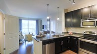 This single-floor residence offers a thoughtfully designed open-concept layout that seamlessly connects the kitchen and living areas, creating a spacious and inviting atmosphere. The kitchen is appointed with rich, dark cabinetry that provides a striking contrast against the light-toned subway tile backsplash and granite countertops. A central kitchen island with an integrated sink and stainless steel dishwasher serves as both a functional workspace and a natural gathering point. Pendant lighting hangs above the island, adding a stylish focal point to the space. The kitchen flows naturally into the living area, which is brightened by large sliding glass doors that invite an abundance of natural light and provide direct access to an outdoor balcony, offering open views of the surrounding landscape. Warm hardwood flooring runs throughout the space, grounding the design and complementing the overall color palette. The living area is well-proportioned, accommodating comfortable seating arrangements alongside a wall-mounted television. The combination of modern finishes, natural light, and the seamless indoor-outdoor connection gives this residence a polished yet welcoming character.