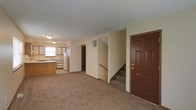 This two-floor residential unit offers a straightforward and functional layout suited for comfortable everyday living. The main level features an open living area with wall-to-wall carpeting that extends throughout, creating a warm and cohesive feel underfoot. A solid entry door with classic six-panel detailing provides a welcoming entryway at the front of the space.

The kitchen is positioned toward the rear of the main level and is equipped with wood-toned cabinetry, a double sink set beneath a window that allows natural light to filter in, and a full-size refrigerator. The cabinetry provides ample storage, and the layout is designed for efficient use of space.

A centrally located staircase with a dark wood handrail connects the main level to the upper floor, offering easy access between the two levels. The staircase is carpeted in keeping with the overall flooring style of the home.

The interior carries a neutral color palette throughout, with cream-toned walls and a textured ceiling finish that adds subtle visual character. Flush-mount ceiling lighting provides consistent illumination across the living areas. The overall ambiance is clean, practical, and move-in ready, making it well-suited for those seeking a no-fuss, functional residential space.