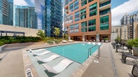 Located in the vibrant Midtown Atlanta neighborhood, this rooftop amenity level at 1081 Juniper St NE offers a thoughtfully designed outdoor retreat set against a dramatic urban skyline backdrop. The centerpiece is a generously sized resort-style swimming pool, lined with contemporary chaise lounges and surrounded by a clean, tiled deck. Adjacent to the pool, a covered cabana lounge area provides a shaded retreat featuring warm-toned paneled walls, a wall-mounted display, and stylish seating arrangements with neutral upholstery and layered rugs, lending an upscale, hospitality-inspired feel. The outdoor terrace extends into multiple distinct zones, including a lush green lawn with oversized white sectional sofas and a slatted wood coffee table, ideal for relaxed social gatherings. Curated garden corridors wind through raised planters filled with diverse flowering shrubs and greenery, creating a tranquil escape from the surrounding cityscape. Additional seating nodes with outdoor dining tables and lounge chairs are anchored around a built-in fireplace feature, offering a cozy gathering spot. Throughout the deck, panoramic views of Atlanta's glittering glass towers frame the space, reinforcing its premier urban setting. The overall ambiance balances sophisticated design with approachable comfort.