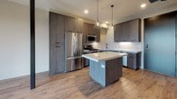 This single-floor residence showcases a thoughtfully designed open layout that balances modern aesthetics with functional living. The kitchen serves as a centerpiece, outfitted with dark wood-grain cabinetry extending floor to ceiling, a full suite of stainless steel appliances including a gas range, built-in microwave, and French door refrigerator. A central island with a light quartz countertop provides additional workspace and is highlighted by glass pendant lighting overhead. Light hardwood flooring flows throughout the main living areas, complementing the neutral color palette.

Distinctive arched windows are a notable architectural feature, flooding the living spaces with generous natural light and offering views of the surrounding neighborhood. One bedroom features plush carpet flooring and a ceiling fan, while another room benefits from a dramatic arched window configuration.

The primary bathroom includes large-format tile flooring, a white undermount sink, and direct access to a walk-in closet with built-in shelving and hanging rods. A second full bathroom features white subway tile surrounding a tub-shower combination, matte black fixtures, and a wood-toned vanity with a light countertop. A balcony extends the living space outdoors, accessible through the main living area.