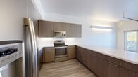 This modern two-bedroom, two-bathroom residence in Denver's 80211 zip code offers a thoughtfully designed single-floor layout. The open-concept kitchen serves as a central feature, showcasing flat-front wood-grain cabinetry extending to the ceiling, stainless steel appliances including a range, microwave, and refrigerator, and a clean white countertop with a peninsula that opens to the living area. Light wood-style flooring flows throughout the main living spaces, enhancing the bright, contemporary aesthetic. The primary bedroom features elevated ceilings, a distinctive glass block window that diffuses natural light, and a ceiling fan. The second bedroom offers a standard window with views of surrounding greenery and shares the same light flooring. Each bathroom is finished with large-format tile flooring and wood-grain vanity cabinetry. The primary bath includes a walk-in shower with white subway tile and a single-sink vanity, while the second bath features a full subway-tiled tub-shower combo. A hallway closet houses a convenient stacked in-unit washer and dryer. Two closets provide ample storage throughout the residence. The overall design presents a clean, modern palette with consistent finishes across all rooms.