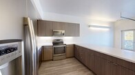 This modern two-bedroom, two-bathroom residence in Denver's 80211 zip code offers a thoughtfully designed single-floor layout. The open-concept kitchen serves as a central feature, showcasing flat-front wood-grain cabinetry extending to the ceiling, stainless steel appliances including a range, microwave, and refrigerator, and a clean white countertop with a peninsula that opens to the living area. Light wood-style flooring flows throughout the main living spaces, enhancing the bright, contemporary aesthetic. The primary bedroom features elevated ceilings, a distinctive glass block window that diffuses natural light, and a ceiling fan. The second bedroom offers a standard window with views of surrounding greenery and shares the same light flooring. Each bathroom is finished with large-format tile flooring and wood-grain vanity cabinetry. The primary bath includes a walk-in shower with white subway tile and a single-sink vanity, while the second bath features a full subway-tiled tub-shower combo. A hallway closet houses a convenient stacked in-unit washer and dryer. Two closets provide ample storage throughout the residence. The overall design presents a clean, modern palette with consistent finishes across all rooms.