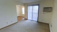 This two-floor residential space offers a practical and functional layout designed for comfortable everyday living. The main living area features wall-to-wall carpeting that adds warmth underfoot, while a large sliding glass door dressed with vertical blinds fills the room with natural light and provides direct access to an outdoor balcony or patio, offering a seamless connection to the outdoors. A wall-mounted HVAC unit ensures climate control convenience. An open archway leads from the living space into an adjacent room finished with hardwood-style flooring, creating a natural flow between spaces. This secondary area benefits from its own window, further enhancing the brightness and airy feel of the interior. Baseboard heating is visible along the walls, providing an efficient supplemental heat source. The neutral, light-toned walls throughout contribute to a clean and open ambiance, making the space easy to personalize. Spanning two floors, the layout offers separation between living and private areas, providing a sense of defined purpose for each level. Overall, the space presents a straightforward, well-lit environment suited for a variety of living arrangements.