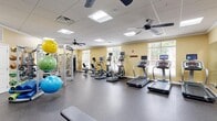 Located at 10449 Atlee Station Rd in Mechanicsville, VA, this fitness center offers a comprehensive workout facility within a single, open-plan floor. The space is designed with functionality and comfort in mind, featuring warm yellow walls, a drop ceiling with recessed panel lighting, and durable rubber flooring throughout. Multiple ceiling fans ensure consistent air circulation across the entire floor.

Natural light fills the room through several large windows fitted with blinds, offering views of the surrounding greenery. A full-length wall mirror visually expands the space and aids users during strength training exercises.

The cardio section is well-stocked with multiple treadmills, elliptical trainers, and stationary recumbent and upright bikes, each equipped with individual media screens for entertainment during workouts. A wall-mounted flat-screen television is also present for shared viewing.

The strength training area includes a multi-station cable machine, a power rack, a dumbbell rack with a variety of free weights, and an assortment of medicine and stability balls. A workout bench provides additional versatility. The thoughtful layout ensures ample open floor space between equipment zones, allowing users to move freely and safely throughout the facility.
