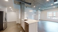 This single-floor unit showcases a striking industrial-modern aesthetic defined by exposed concrete ceilings with raw, textured finishes and polished concrete floors that run throughout the space. Structural concrete columns add an authentic loft character, while crisp white walls provide a clean contrast throughout.

The open-concept kitchen is a central highlight, featuring a large waterfall-edge island with an undermount sink, white lower cabinetry, and dark espresso upper cabinets. Stainless steel appliances — including a gas range, built-in microwave, and French door refrigerator — complete the kitchen's modern and functional design. Pendant lights hang above the island, complementing the recessed lighting overhead.

The living area flows seamlessly from the kitchen, benefiting from soaring ceiling heights that amplify the sense of openness. Bedrooms are generously proportioned with clean white walls and polished concrete floors; one bedroom features a large window offering views of the surrounding neighborhood and greenery, filling the room with natural light.

The bathrooms maintain the same clean, modern palette, featuring white cabinetry with chrome hardware, undermount sinks, and full tub-shower combinations clad in white subway tile. The polished concrete flooring continues here as well, reinforcing the unit's cohesive industrial character throughout every room.