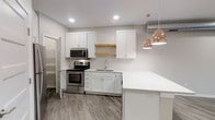 This single-floor space showcases a thoughtfully designed kitchen that balances modern aesthetics with functional details. White shaker-style cabinetry lines the walls, complemented by a classic white subway tile backsplash that adds texture without overwhelming the clean palette. A natural wood floating shelf introduces a warm contrast between the upper and lower cabinets, providing both display space and visual interest.

Stainless steel appliances, including an electric range and over-the-range microwave, offer a sleek, contemporary finish. The undermount sink is neatly integrated into the lower cabinetry, maintaining the streamlined look. A generous kitchen peninsula with a light quartz countertop extends the workspace and creates a natural division between the kitchen and the adjoining area.

Rose gold dome pendant lights hang above the peninsula, adding a touch of warmth and elegance to the space. Recessed lighting throughout ensures the area is well-lit. The wood-look flooring in a neutral gray tone runs throughout, tying the space together cohesively. A partially open door hints at additional storage or utility space nearby, adding to the overall practicality of the layout.