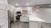 This single-floor space showcases a thoughtfully designed kitchen that balances modern aesthetics with functional details. White shaker-style cabinetry lines the walls, complemented by a classic white subway tile backsplash that adds texture without overwhelming the clean palette. A natural wood floating shelf introduces a warm contrast between the upper and lower cabinets, providing both display space and visual interest.

Stainless steel appliances, including an electric range and over-the-range microwave, offer a sleek, contemporary finish. The undermount sink is neatly integrated into the lower cabinetry, maintaining the streamlined look. A generous kitchen peninsula with a light quartz countertop extends the workspace and creates a natural division between the kitchen and the adjoining area.

Rose gold dome pendant lights hang above the peninsula, adding a touch of warmth and elegance to the space. Recessed lighting throughout ensures the area is well-lit. The wood-look flooring in a neutral gray tone runs throughout, tying the space together cohesively. A partially open door hints at additional storage or utility space nearby, adding to the overall practicality of the layout.