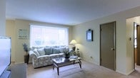 This single-floor space offers a comfortable and inviting layout designed for relaxed everyday living. The living room serves as the central gathering area, showcasing soft, wall-to-wall carpeting that adds warmth underfoot and contributes to the room's cozy atmosphere. A large picture window dressed with vertical blinds allows generous natural light to fill the space, illuminating the neutral, light-toned walls that create a clean and airy feel. The room comfortably accommodates a full-sized sofa complemented by decorative throw pillows, centered around a glass-top coffee table — a functional yet stylish focal point. A classic table lamp on an end table provides supplementary ambient lighting, enhancing the room's welcoming character. A decorative wall-mounted mirror adds a charming accent to the otherwise understated wall space. A doorway visible from the living area suggests easy access to additional rooms, indicating a practical and well-connected floor plan. Overall, the space exudes a calm, livable quality with its soft color palette, natural light, and unpretentious furnishings — well-suited for those seeking a straightforward and comfortable living environment.