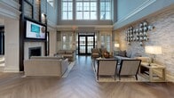 Sutton Landing is an elegantly designed two-floor amenity space located at 300 Sutton Court in Mount Sinai, New York. The facility greets visitors with a stunning entryway showcasing herringbone hardwood floors, wainscoting, and a dramatic staircase featuring dark treads, glass panels, and gold brass railings — anchored by a striking grand piano that sets a refined tone throughout.

The main lounge area offers a warm and inviting atmosphere centered around a stacked-stone fireplace with a mounted television above, flanked by comfortable seating arrangements. Adjacent dining and social areas feature coffered dark wood ceilings, neutral tones, and curated artwork, creating an upscale yet welcoming environment.

The game room is a standout space, outfitted with a billiards table, bar-height seating, a projection screen, and dramatic dark millwork with gold-accented coffered ceilings. A well-appointed kitchenette with modern cabinetry, white quartz countertops, and subway tile backsplash provides convenient service support.

The fitness center is equipped with a full array of cardio and strength machines, plantation shutters allowing ample natural light, and a wall-mounted television. Elevator access connects both floors seamlessly, ensuring full accessibility throughout the facility.