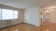 This single-floor space features a clean and minimalist aesthetic highlighted by warm hardwood flooring that runs throughout the main living area. The open layout creates a sense of spaciousness, with smooth white walls and a standard ceiling height that contribute to a bright, airy feel. A window along one wall allows natural light to filter into the living area, illuminating the space and enhancing its understated charm. Baseboard heating units line the perimeter of the room, providing an efficient and unobtrusive heating solution. A partial wall divides the main living area from an adjacent hallway that leads to the kitchen, which is visible in the background and features warm wood cabinetry. The transition between rooms is seamless, with the consistent hardwood flooring tying the spaces together cohesively. The overall ambiance is neutral and versatile, offering a blank canvas well-suited for a variety of interior design approaches. The simplicity of the finishes and the functional layout make this a practical and adaptable space.