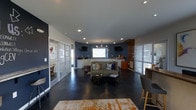 This single-floor space offers an open-concept layout that seamlessly blends a lounge area with a kitchenette, creating a versatile and functional environment. The polished concrete floors lend an industrial-chic aesthetic, grounding the space with a durable, low-maintenance surface that complements the overall design.

The focal point of the room is a bold navy patterned accent wall that anchors the kitchenette area, which features white cabinetry, a countertop workspace, and warm wood accents. A central window brings in natural light, brightening this area considerably. Two wall-mounted flat-screen TVs flank the kitchenette, adding a media-ready functionality to the space.

The lounge area is furnished with a green sofa accented by colorful throw pillows, centered around a low coffee table. A warm-toned area rug adds texture and definition to the seating zone. Recessed lighting throughout the ceiling is complemented by a statement chandelier, providing layered illumination.

On either side of the main space, frosted glass-paneled doors lead to adjoining rooms, offering privacy while maintaining an open, airy feel. A bar-height counter with seating is positioned to one side, adding a social gathering point.