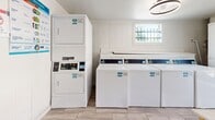 Located at 910 Fair Oaks Rd in Houston, TX, this shared laundry facility offers a clean, efficiently organized space designed to meet residents' laundering needs. The room features wood-look tile flooring throughout, white panel walls, and a modern circular ceiling light fixture that provides bright, even illumination across the entire area.

The laundry room is well-equipped with multiple front-row top-load washing machines and a stacked washer-dryer unit, all finished in white and clearly labeled for easy use. A row of top-load washers lines one wall, while a stacked dryer unit maximizes vertical space in the corner. A central folding table with a wood frame and marble-style surface provides a convenient workspace for sorting and folding laundry.

Natural light filters in through a barred window, adding to the bright and airy feel of the space. A secured iron-barred door with a full-length window provides the main point of entry, offering both ventilation and a view of the outdoors. A wall-mounted ventilation grille and a fire extinguisher are also present, reflecting attention to safety and airflow. Informational signage near the machines guides users through the laundry process, making this a functional and user-friendly shared amenity.
