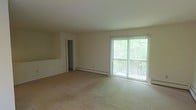 This single-floor residence offers a clean, functional layout designed to maximize comfort and usability. The living area is generously proportioned, featuring soft wall-to-wall carpeting underfoot and neutral-toned walls that create a warm, welcoming atmosphere. A large sliding glass door floods the space with abundant natural light and provides direct access to a private balcony overlooking a verdant, tree-lined setting — an ideal spot to enjoy the outdoors. A baseboard heating system runs along the exterior wall, ensuring efficient climate control throughout the cooler months. An interior doorway leads to additional rooms, offering a well-connected flow throughout the unit. The overall design is straightforward and practical, with a light color palette that enhances the sense of openness and makes the space easy to personalize. The combination of ample natural light, outdoor access, and open living space gives this residence a relaxed, airy ambiance well-suited for everyday living.