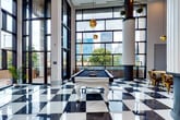 Located at 101 W Morehead St in Charlotte, NC, this striking amenity lounge delivers a one-of-a-kind experience through its dramatic architectural design and vibrant, curated interiors. The space is anchored by gleaming black-and-white marble checkerboard flooring and soaring double-height ceilings that amplify the sense of grandeur throughout.

Floor-to-ceiling windows flood the interior with abundant natural light while framing sweeping views of the Charlotte skyline and an outdoor pool area. Towering fluted columns add a classical architectural counterpoint to the space's eclectic, contemporary aesthetic.

The lounge is thoughtfully divided into distinct functional areas. A game area features a statement white billiards table set against the backdrop of the city skyline. A communal dining area is outfitted with a rustic wood table, gold metal chairs, and warm pendant lighting. An integrated kitchenette showcases pink subway tile accents, white countertops, and stainless steel appliances.

A curated reading nook houses a built-in bookshelf stocked with games and literature. Seating vignettes throughout the space feature ornate black velvet chairs, gold accent tables, and textured wall coverings. A bold gallery wall displaying gold-framed pop art portraits serves as a focal artistic centerpiece, reinforcing the space's dynamic, culturally rich character.