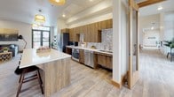 This single-story community clubhouse is thoughtfully designed to provide residents with a welcoming and functional gathering space. The interior design blends warm wood tones, light-colored luxury vinyl plank and tile flooring, and crisp white walls to create a modern yet approachable aesthetic throughout.

The main social area features a comfortable lounge with a gray sectional sofa, layered area rugs, and large windows that invite natural light and views of the surrounding neighborhood. Landscape-inspired artwork and lush indoor plants add warmth and personality to the space.

The fully equipped kitchen is a standout feature, showcasing medium-toned wood cabinetry, stainless steel appliances including a refrigerator, range, and dishwasher, white quartz countertops, and a decorative mosaic tile backsplash. A large kitchen island provides additional counter space and casual seating.

Adjacent to the kitchen, a dedicated dining area accommodates multiple tables with black metal chairs, ideal for group gatherings. A pass-through window with warm wood trim connects to the lounge area, enhancing the open flow. A built-in coffee station nearby adds everyday convenience.

The game room, crowned by vaulted ceilings and pendant lighting, centers around a full-size pool table and includes a foosball table and wall-mounted television, making it an engaging entertainment destination.
