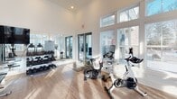 This single-level fitness center is designed with both functionality and aesthetics in mind, offering a bright, open environment ideal for a full range of workout activities. The space features soaring ceilings with elegant coffered and shiplap detailing, complemented by recessed lighting and a ceiling fan that enhance ventilation and comfort throughout. Large, multi-pane windows line the walls, flooding the interior with abundant natural light and providing views of the surrounding exterior. Light hardwood-style flooring runs throughout, contributing to the clean, modern atmosphere.

The cardio zone is well-appointed with a variety of equipment, including treadmills, elliptical trainers, rowing machines, and spinning bikes, arranged to maximize both space and usability. The strength training area features a Precor functional training system, a Smith machine with free weights, a dumbbell rack, and additional resistance machines, catering to a wide range of fitness goals. Full-length mirrors along one wall visually expand the space while supporting proper exercise form. Glass doors open to an outdoor area, further enhancing the sense of openness. Overall, the space presents a polished, professional fitness environment suited for comprehensive daily training.