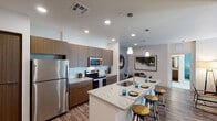 Located at 5151 N 95th Ave in Glendale, Arizona, this contemporary single-story residence offers a thoughtfully designed open-concept layout finished with modern fixtures and warm, neutral tones throughout. The kitchen is a standout feature, boasting flat-front wood-tone cabinetry, a white subway tile backsplash, stainless steel appliances including a refrigerator, electric range, and built-in microwave, and a large granite-topped island with an undermount sink and bar seating for multiple guests, illuminated by sleek pendant lighting. Wood-look flooring flows through the main living areas, connecting the kitchen to a dedicated dining nook and a comfortable living room accented by a bold teal feature wall, ceiling fan, and glass doors that open to an outdoor area. The primary bedroom features plush carpet, a ceiling fan, a window for natural light, and access to a private bathroom with granite countertops, wood-tone cabinetry, a combined tub and shower, and an adjoining walk-in closet. A second bedroom, also carpeted and ceiling fan-equipped with its own window, is served by a second full bathroom with matching finishes and a walk-in shower. Recessed lighting and clean white walls create a bright, cohesive ambiance throughout the entire space.