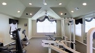 This single-floor fitness room offers a well-appointed space designed for home workouts. The most notable architectural feature is the vaulted ceiling, which rises to a central peak and is finished in a soft sage green tone, creating an open, airy atmosphere. Recessed lighting is strategically placed throughout the angled ceiling panels, ensuring bright, even illumination across the room. A ceiling fan is centrally mounted, providing consistent airflow during exercise sessions.

The room is equipped with a variety of fitness equipment, including a multi-station cable weight machine, an adjustable weight bench, and an upright stationary bike, offering a well-rounded range of workout options. The flooring consists of wall-to-wall carpeting in a neutral tone, providing cushioning underfoot.

A window centered on the far wall is dressed with decorative dark fabric valances and white horizontal blinds, allowing natural light to filter in while maintaining privacy. The walls are painted in a warm neutral beige, complementing the overall calm and functional ambiance of the space.