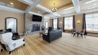 Located on Offshore Drive in East Amherst, NY, this single-floor community clubhouse offers a well-appointed gathering space designed with both comfort and functionality in mind. The architecture is defined by signature coffered ceilings with white trim detail throughout the main areas, pendant chandeliers, and warm neutral tones that create a welcoming, upscale atmosphere.

The entrance foyer sets an immediate tone with large-format tile flooring, bold patterned wallcovering, recessed accent lighting, and a pair of leather armchairs flanking a decorative mirror. French doors with glass panels provide a bright transition into the interior.

The main lounge area is a standout, anchored by a floor-to-ceiling stacked stone fireplace with a wall-mounted television above. Leather seating and accent chairs are arranged for comfort, while the coffered ceiling adds architectural depth. Natural light streams through multiple windows dressed with decorative patterned drapery.

Adjacent to the lounge, a wet bar area features a curved dark wood counter with granite top, bar stools, and a mounted television — ideal for entertaining. A separate full kitchen includes rich wood cabinetry, granite countertops, stainless steel appliances, and tile flooring. Glass-paneled double doors provide outdoor access, completing this versatile community space.