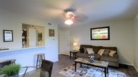 This single-floor space is designed with an open, functional layout that seamlessly connects the living, dining, and kitchen areas. The living room serves as the central gathering space, featuring wood-style laminate flooring that runs throughout, adding warmth and visual continuity. A ceiling fan with a built-in light fixture is centrally mounted, providing both airflow and ambient lighting. The living area comfortably accommodates a sofa with decorative pillows, a granite-top coffee table, and a patterned area rug that anchors the seating arrangement.

Adjacent to the living area, a pass-through counter with bar seating connects the kitchen to the main living space, creating an inclusive, social atmosphere. A barstool and dining chair are visible near this counter, offering casual seating options. The entryway is positioned at the far end of the living room, marked by a front door flanked by neutral walls. A framed mirror near the entry reflects the living space, adding depth to the room. Overall, the space maintains a neutral color palette with white walls and warm flooring, creating a clean, inviting atmosphere throughout.