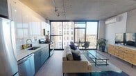 Located in Chicago's Uptown neighborhood, this single-floor unit at 4555 N Sheridan Road showcases a sleek, modern design defined by its signature exposed concrete ceilings and light hardwood flooring. The open-concept living and kitchen area is anchored by dramatic floor-to-ceiling windows that wrap the corner, flooding the space with natural light and offering expansive views of the Chicago skyline and Lake Michigan beyond.

The kitchen is outfitted with stainless steel appliances — including a French-door refrigerator, gas range, and built-in microwave — complemented by dual-tone cabinetry featuring white upper cabinets and blue-grey lower cabinets, a classic subway tile backsplash, and a stainless steel sink. The living area provides generous room for lounging and dining, with a glass-top coffee table and a dedicated dining nook positioned directly at the windows to take full advantage of the views.

The bedroom is accessible via a sliding frosted-glass panel, offering privacy while maintaining the unit's open aesthetic. A wall-mounted mini-split system provides climate control throughout. The bathroom features clean, contemporary finishes with dark tile flooring, a floating vanity, and a large mirror. Track lighting and recessed fixtures add to the modern industrial ambiance of this well-appointed urban residence.