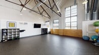 This single-floor fitness and activity studio is defined by its impressive architectural character and open, functional layout. The space is anchored by soaring vaulted ceilings with exposed dark steel trusses and white-painted rafters, lending it an industrial yet refined aesthetic. Large, multi-pane industrial windows line the walls, flooding the interior with abundant natural light and offering views of the surrounding urban environment.

The lower walls feature a distinctive warm-toned yellow brick wainscoting, which contrasts handsomely with the crisp white upper walls. The flooring throughout consists of durable dark rubber/epoxy material, well-suited for high-activity use. Exposed ductwork and pendant lighting fixtures run along the ceiling, reinforcing the space's industrial character.

The main open area is generously proportioned, offering ample room for group fitness classes, training sessions, or flexible programming. A wall-mounted flat-screen TV and built-in speakers support instructional or media needs. Exercise equipment including medicine balls and yoga accessories is neatly stored along one wall. A modular frosted-glass partition system at one end provides the option to subdivide the space. The overall ambiance is energetic and versatile, blending historic architectural elements with modern fitness infrastructure.